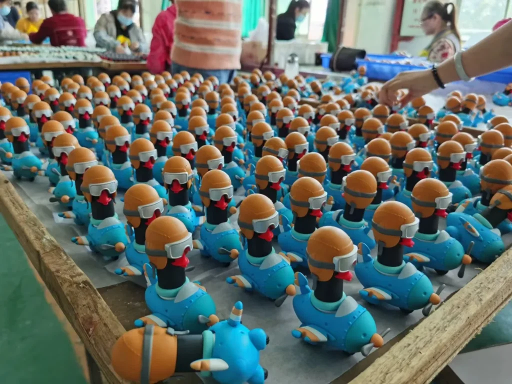 Rows of blue vinyl duck toys wearing helmets on an assembly line with workers in the background at a toy factory.