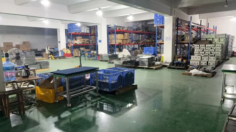 Spacious industrial warehouse with metal racks, stacked cardboard boxes, and blue crates on green flooring, under bright lighting.