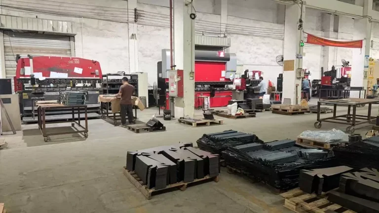 Interior of a metal fabrication workshop featuring large machinery, metal sheets, and workers handling equipment and materials.