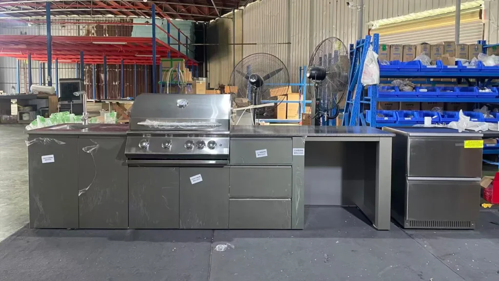 Stainless steel outdoor kitchen setup with cabinets and grill in a workshop with shelving and industrial fans.