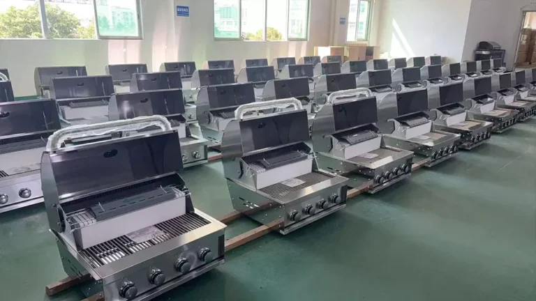 Multiple gas barbecues aligned in rows inside a factory, each with a metal finish and control knobs, on a green floor.
