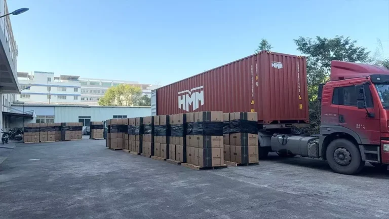 A red truck with an HMM shipping container parked in a logistics area, surrounded by stacked cardboard boxes ready for shipment.